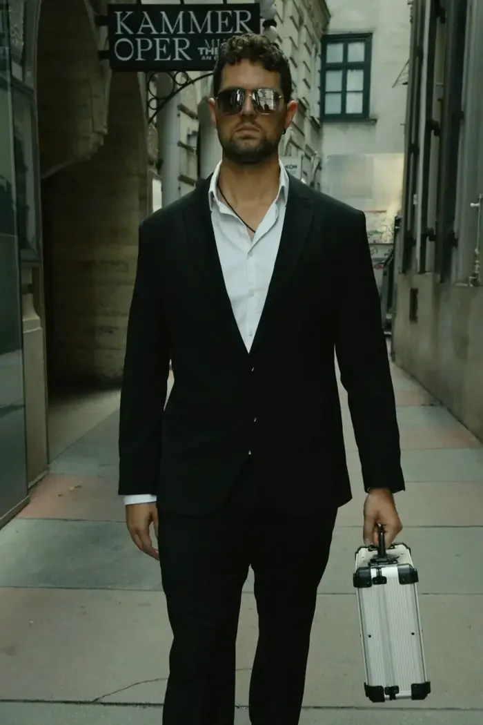 An actor in a dark suit and sunglasses carrying an aluminium case past the Kammeroper in Vienna